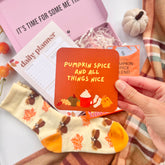 Hand holding a 'Pumpkin Spice and All Things Nice' coaster with fall-themed items like socks and a planner in the background.